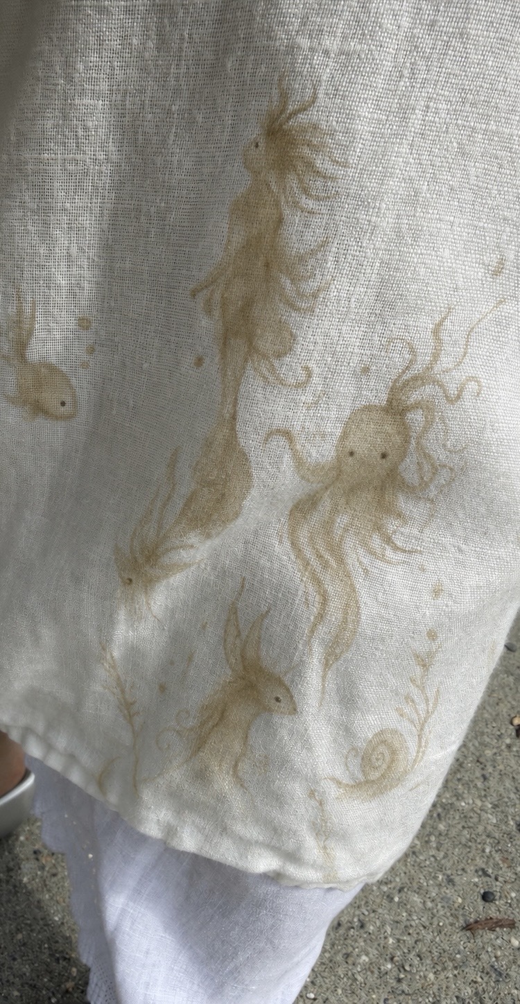 White linen, hem, 17 July 2026. Record of a momentary connection surviving repeated laundering. The field, unable to express itself from within itself, has been written upon by the generative force: a fish at upper left, an octopus at centre right, a spiral shell at lower right, fronds and drifting particulate throughout. No figure was applied by the author. The marks are the law's own handwriting.
